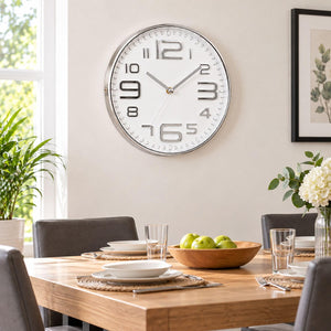 A round wall clock with a silver frame and white face hangs on a beige wall above a wooden dining table set with plates, glasses, and a bowl of green apples. Gray chairs, a potted plant, and framed wall art are visible in the room.