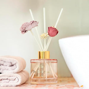 Reed diffuser with floral elements on a wooden surface with towels and a lamp in the background