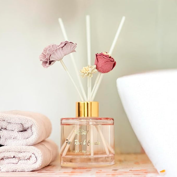 Reed diffuser with floral elements on a wooden surface with towels and a lamp in the background