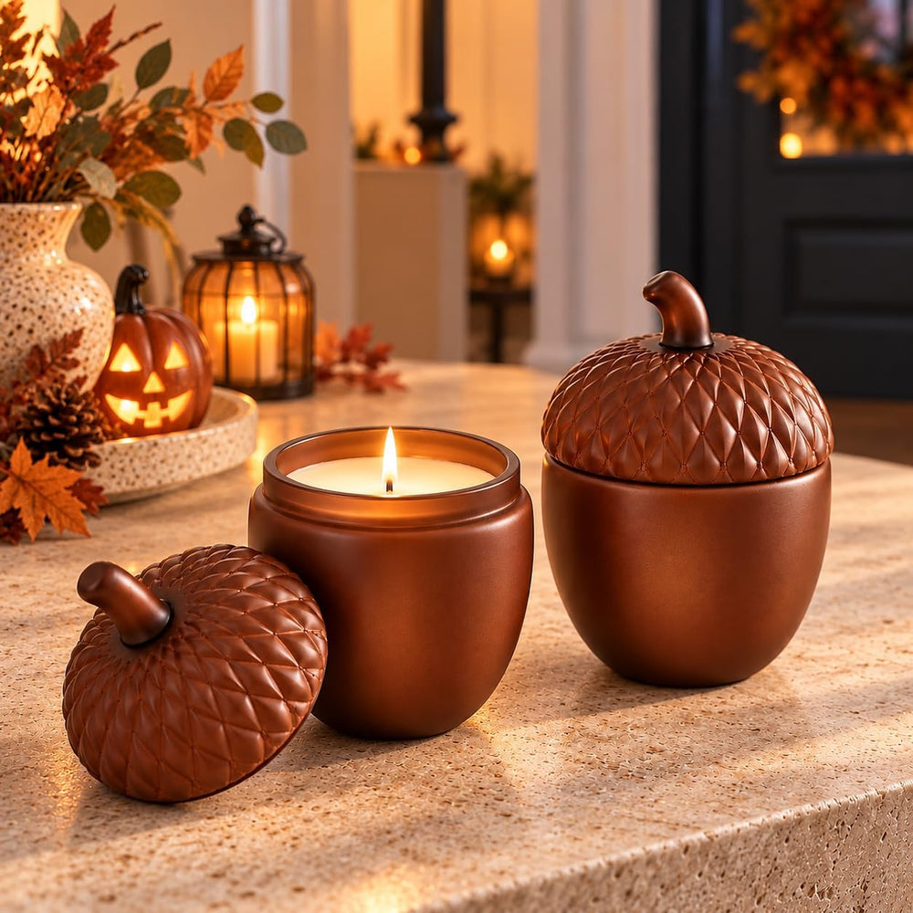 Two acorn-shaped candles in a warm brown ceramic finish, one open with a lit candle inside and one closed, displayed on a stone countertop in a cozy fall-themed setting.