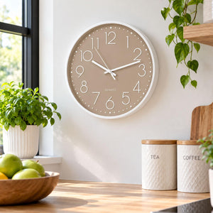 A round wall clock with a white frame and a warm taupe-beige face hangs on a white wall in a bright kitchen setting. The clock features white hands and black Arabic numerals.