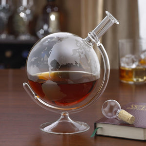 Globe-shaped glass with liquid on a wooden surface, surrounded by other glasses and a book.