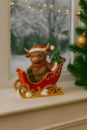 Decorative figurine of a Highland cow in a sleigh with a Christmas tree, on a surface with a blurred background.