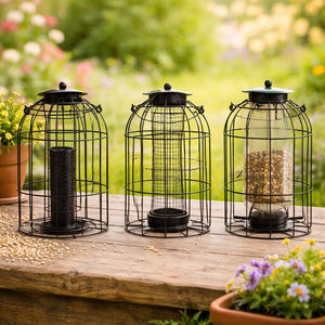 Three black metal bird feeder cages with clear glass jars are arranged on a wooden table in a garden setting. One feeder is empty, and the other two contain birdseed.