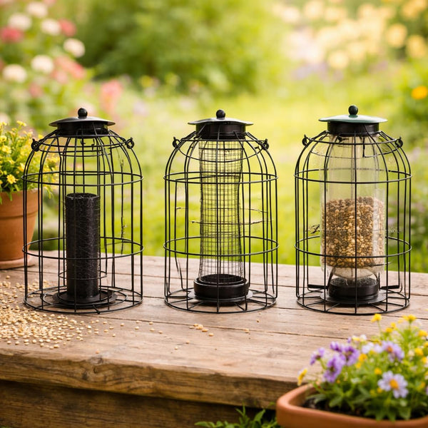 Three black metal bird feeder cages with clear glass jars are arranged on a wooden table in a garden setting. One feeder is empty, and the other two contain birdseed.