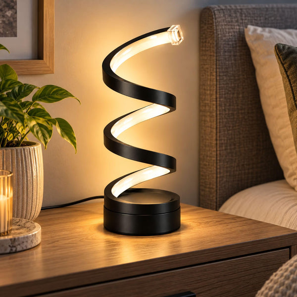 A modern spiral-shaped LED night light with a matte black finish and warm white glow, placed on a wooden bedside table in a bedroom setting next to a plant and bed headboard.