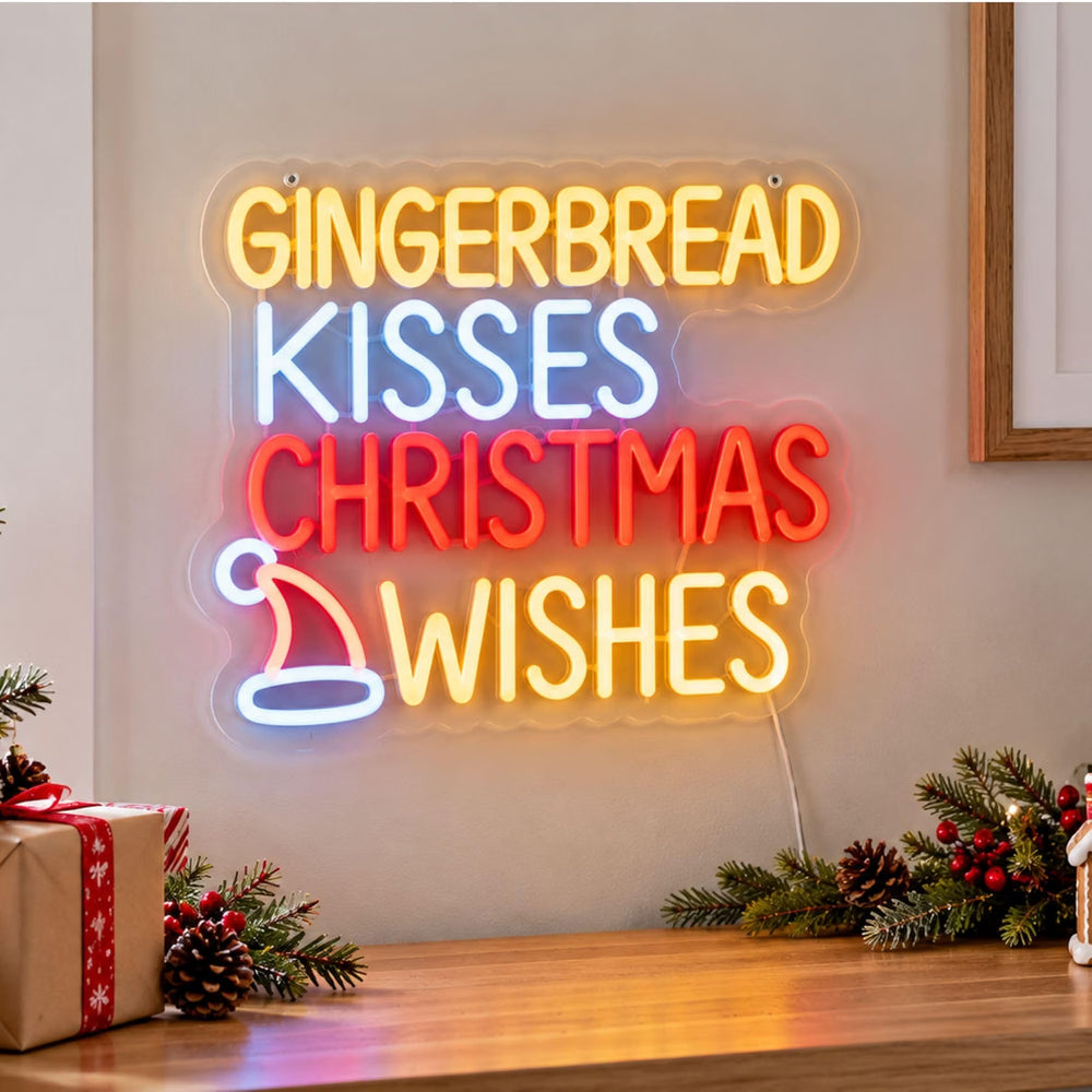 A glowing LED neon-style wall sign with the phrase “GINGERBREAD KISSES CHRISTMAS WISHES” in yellow, blue, and red letters, mounted on a beige wall above a wooden table with holiday decorations.