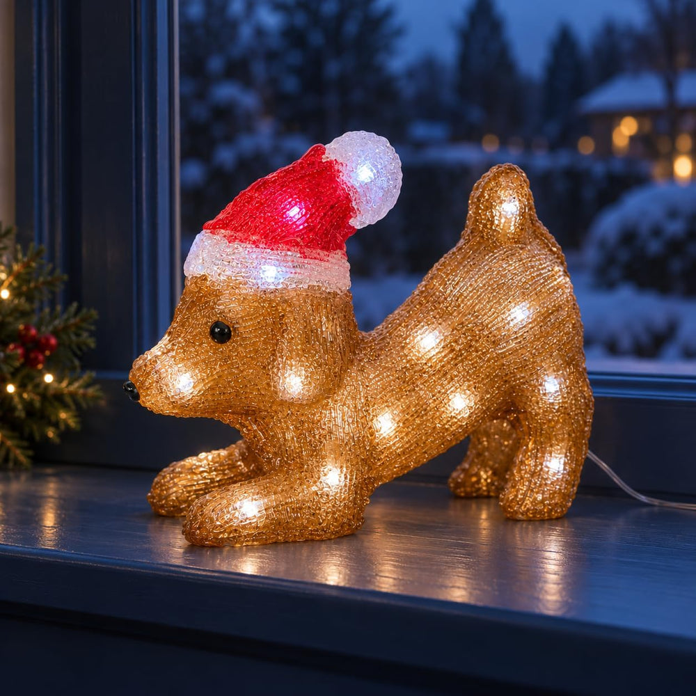 A glittery golden dog-shaped holiday light decoration wearing a red Santa hat with a white pom-pom, posed on a dark wooden surface in front of a softly blurred festive doorway.