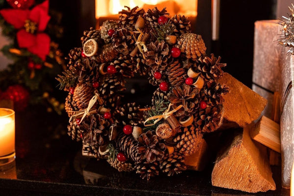 Decorative wreath made of pine cones and berries on a fireplace mantle with candles and logs.