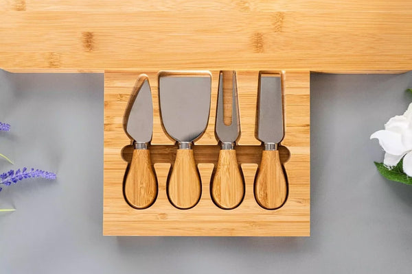 Four-piece cheese and butter knife set with light wood handles and stainless steel blades, presented in a matching wooden tray on a gray surface with decorative flowers.