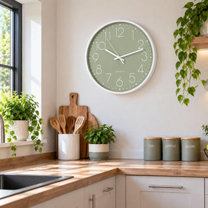 A round wall clock with a sage green face and white frame hangs on a light beige wall in a bright, modern kitchen. The kitchen features wooden countertops, white cabinets, green potted plants, and three gray canisters labeled TEA, COFFEE, and SUGAR.