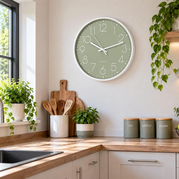 A round wall clock with a sage green face and white frame hangs on a light beige wall in a bright, modern kitchen. The kitchen features wooden countertops, white cabinets, green potted plants, and three gray canisters labeled TEA, COFFEE, and SUGAR.
