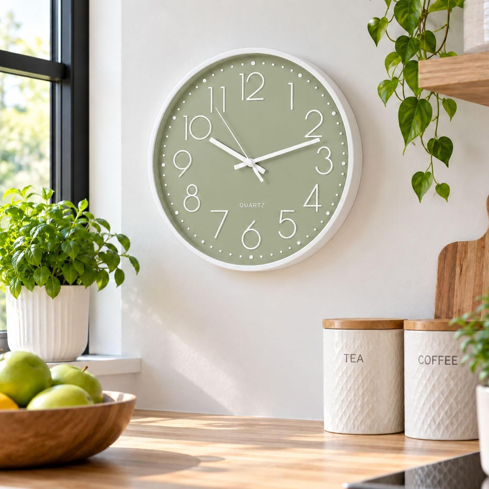 A round wall clock with a sage green face and white frame hangs on a light wall above a wooden countertop. The scene includes potted green plants, white canisters labeled TEA and COFFEE, and a bowl of green apples, creating a bright kitchen or cafe-like interior setting.