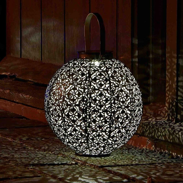A spherical metal lantern with an intricate floral cutout pattern, glowing with warm light and casting shadows on a dark wooden surface and background.