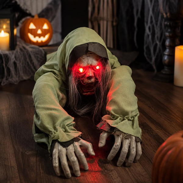 A creepy Halloween decoration of a crawling zombie or ghost in a tattered green hoodie, with glowing red LED eyes and skeletal hands, positioned on a wooden floor in a spooky room setting with candles and a jack-o'-lantern.