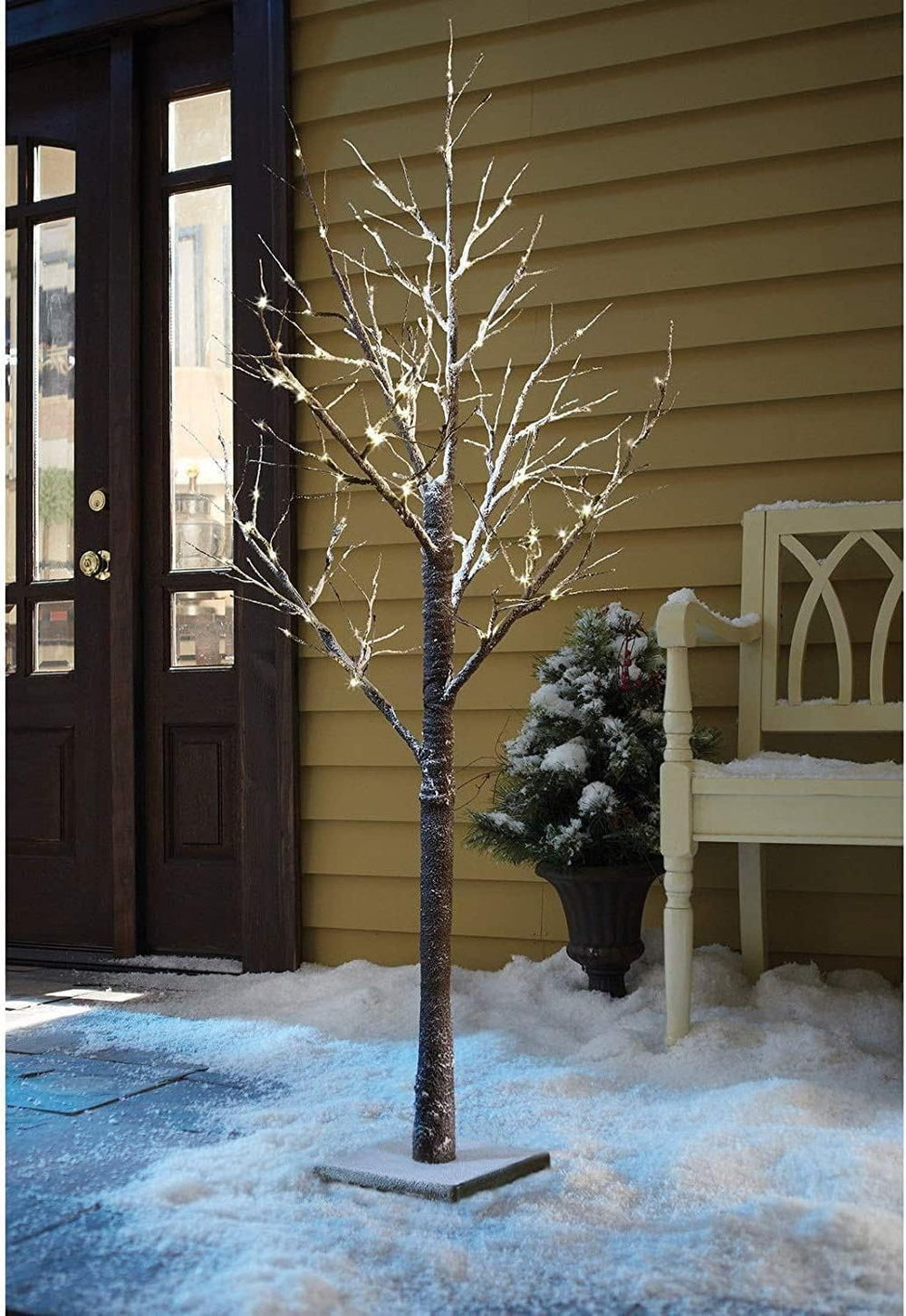 Decorative lighted tree on a snowy porch with a bench and door in the background.