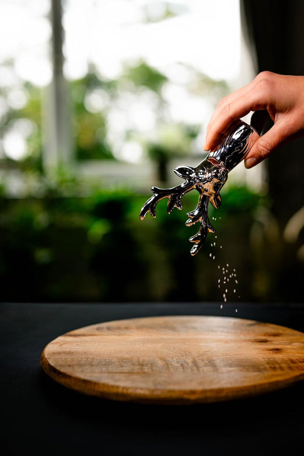 Hand sprinkling spices from a black pepper grinder onto a wooden surface with a blurred natural background