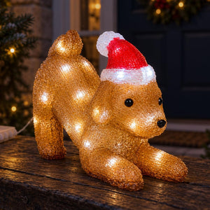 A glittery golden dog-shaped holiday light decoration wearing a red Santa hat with a white pom-pom, posed on a dark wooden surface in front of a softly blurred festive doorway.