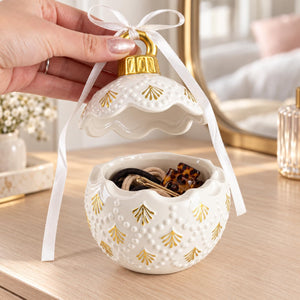 A hand holds open a white ceramic jewelry box with a lid shaped like a scalloped ornament, decorated with gold pinecone motifs and a white ribbon loop. The box is filled with hair ties and clips on a light wood vanity surface.
