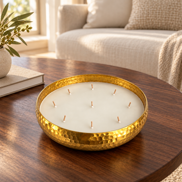 A hammered gold metal candle holder with multiple wicks, filled with white wax, set on a dark wooden table in a cozy living room setting.