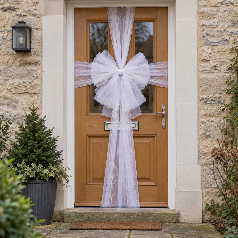 A large white tulle bow and matching hanging tulle decoration displayed on a wooden front door, with a stone wall, wall lantern, potted plants, and a doormat visible.