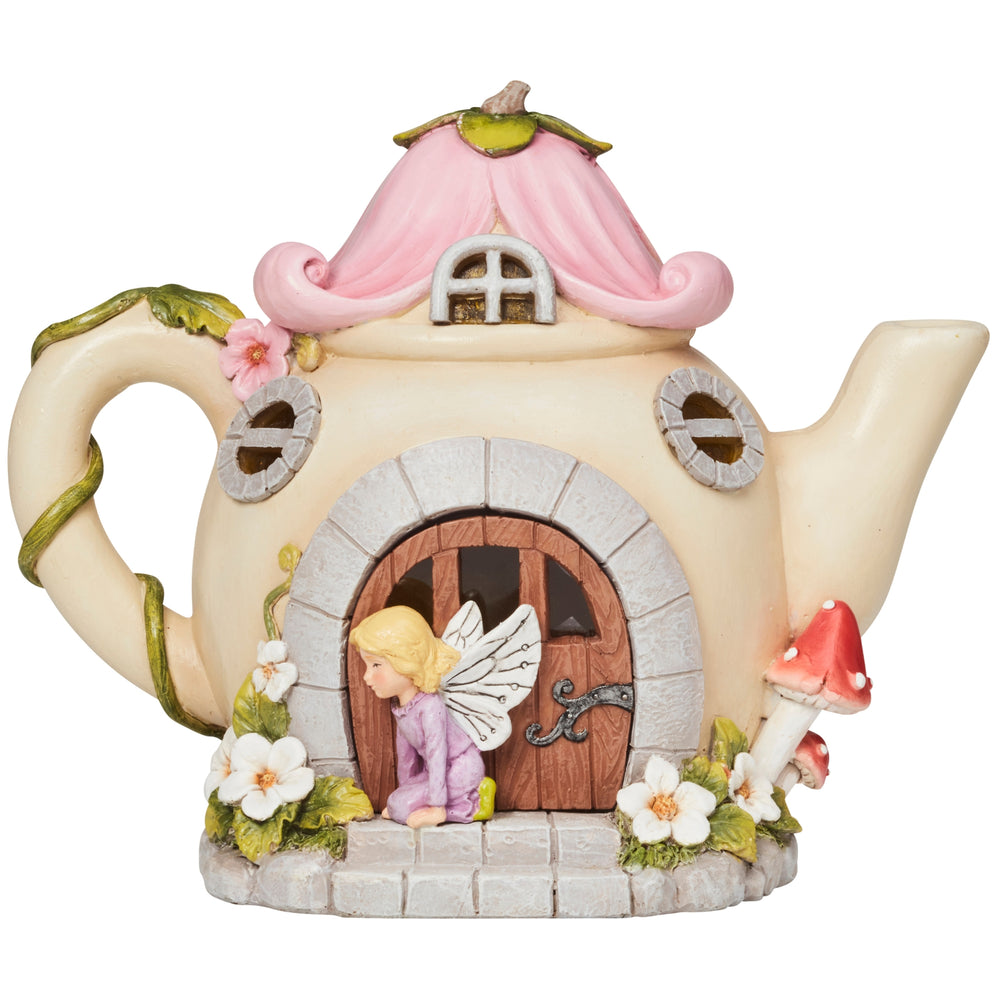 A decorative teapot-shaped fairy house ornament with a pink roof, glowing warm lights, a stone-textured base, and a small winged fairy figure in front of the door. The ornament is styled on a rustic surface with autumn leaves and pumpkins in the background.
