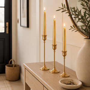 Three tall brass candlesticks with lit taper candles are arranged on a light wood console table in a styled interior setting.