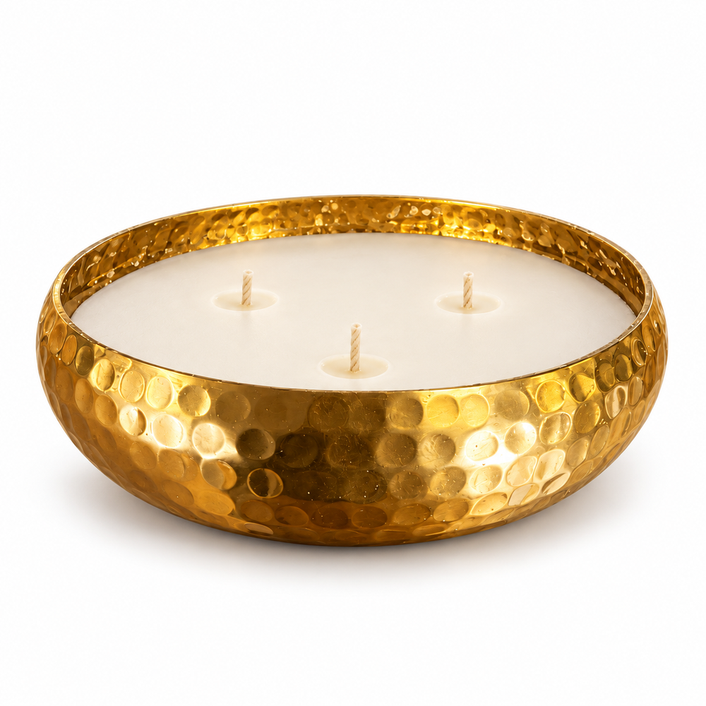 A hammered gold metal candle holder containing three lit white candles, placed in a warm toned living room.