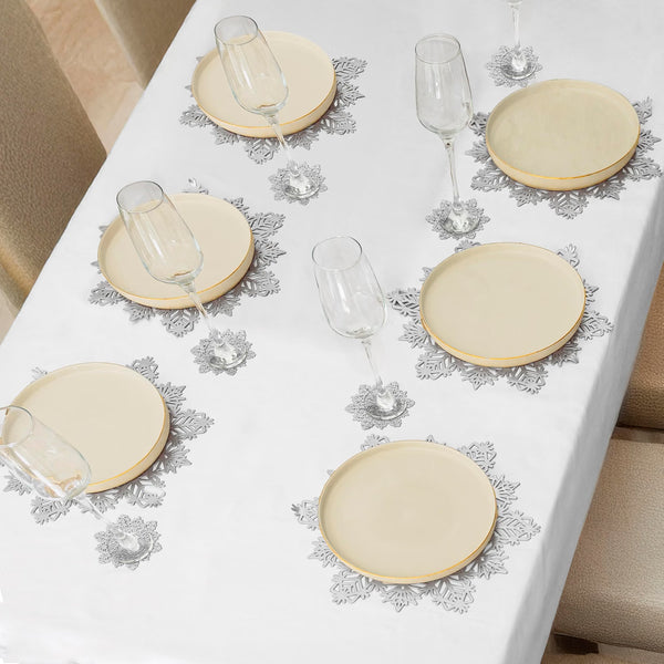 A white tablecloth with a delicate gray floral lace pattern, styled with round cream-colored plates and clear wine glasses on a dining table.