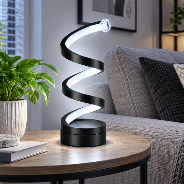 A modern spiral LED table lamp with a black base and illuminated white curve, placed on a round wooden table in a living room setting next to a potted plant and sofa.