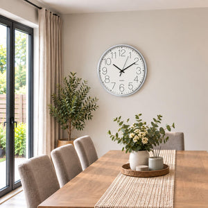 A round wall clock with a white face and silver frame hangs on a beige wall above a wooden dining table with chairs, plants, and a tray of flowers in a bright dining room.
