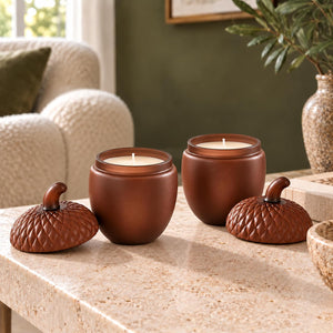 Three brown ceramic candle jars with matching acorn-shaped lids sit on a stone table in a cozy living room setting.