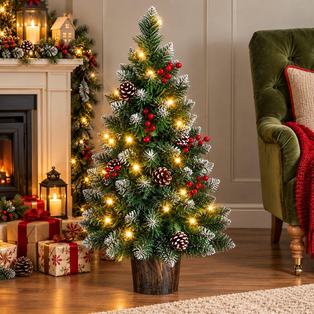 A small artificial Christmas tree with green needles, white snow-like dusting, warm white string lights, red berries, and pinecones, displayed in a cozy living room next to a fireplace and green armchair.