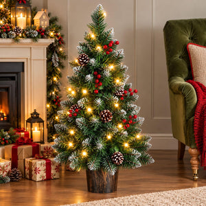 A small artificial Christmas tree with green needles, white snow-like dusting, warm white string lights, red berries, and pinecones, displayed in a cozy living room next to a fireplace and green armchair.