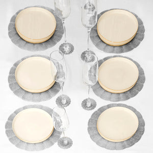 Six place settings arranged on a white tablecloth, each featuring a round cream plate with a gold rim over a gray scalloped placemat, with two clear stemmed glasses beside each setting.