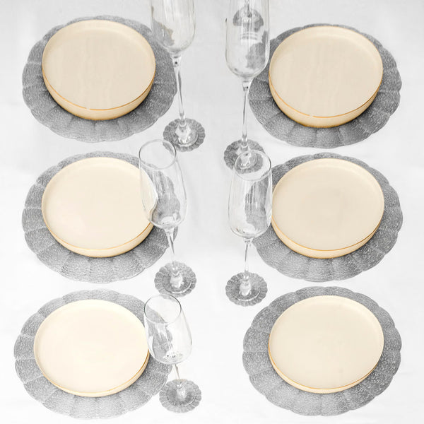 Six place settings arranged on a white tablecloth, each featuring a round cream plate with a gold rim over a gray scalloped placemat, with two clear stemmed glasses beside each setting.