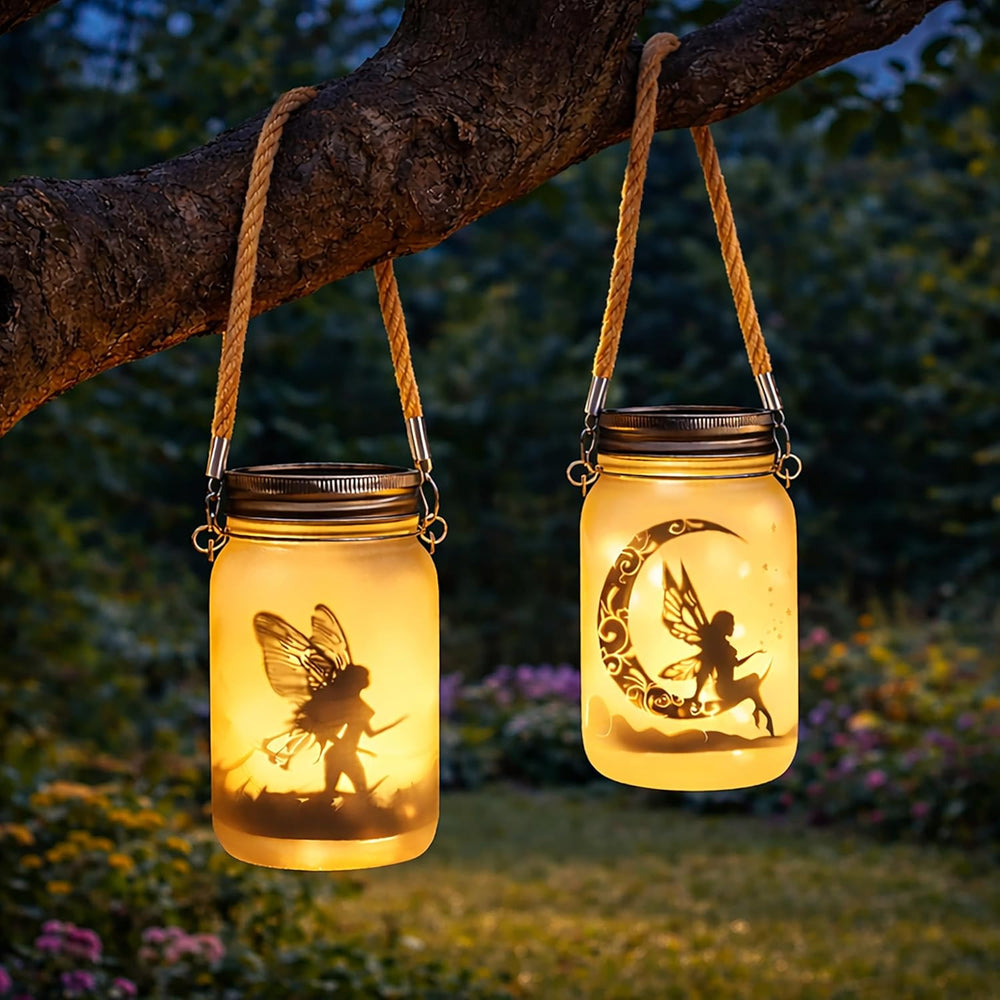 Two mason jar lanterns with fairy and moon designs hanging from a branch at night.