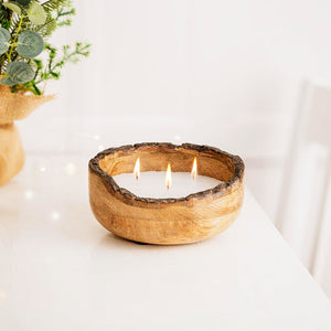 A three-wick soy wax candle in a rustic, handcrafted wooden bowl with a natural bark edge, lit with three small flames on a white tabletop.