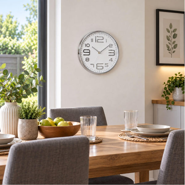 A round wall clock with a white face and silver frame hangs on a light beige wall above a wooden dining table set with gray chairs, glassware, and potted plants in a bright, modern dining room.
