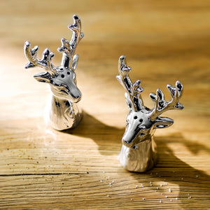 Two small metallic deer head figurines with antlers, photographed on a warm wooden surface in bright sunlight.
