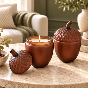 Three amber-brown acorn-shaped candle holders with textured scales and stems, displayed on a stone table in a softly lit living room. One holder is open with a lit candle inside, and two are closed, showing the matching lids.
