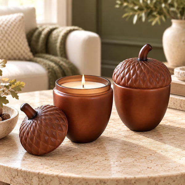 Three amber-brown acorn-shaped candle holders with textured scales and stems, displayed on a stone table in a softly lit living room. One holder is open with a lit candle inside, and two are closed, showing the matching lids.