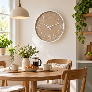 A round wall clock with a beige face and white frame hangs above a wooden dining table in a bright, modern kitchen or dining room. The clock has large white numbers and simple black hands.