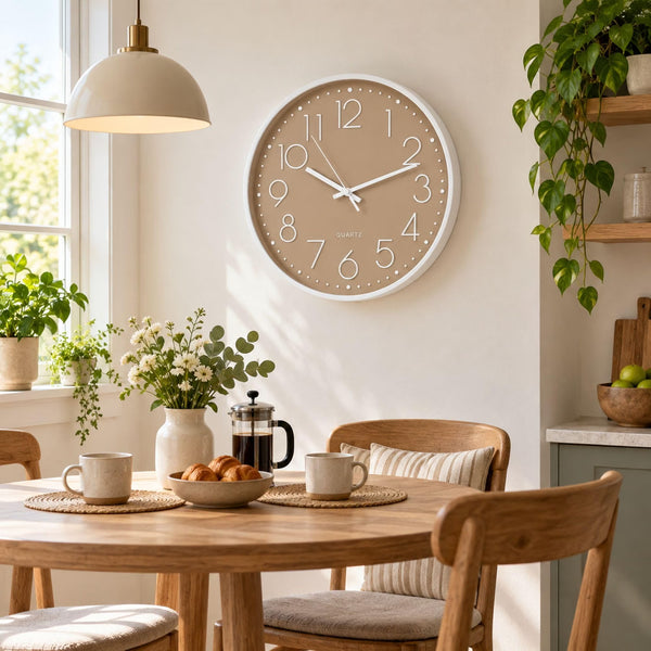 A round wall clock with a beige face and white frame hangs above a wooden dining table in a bright, modern kitchen or dining room. The clock has large white numbers and simple black hands.