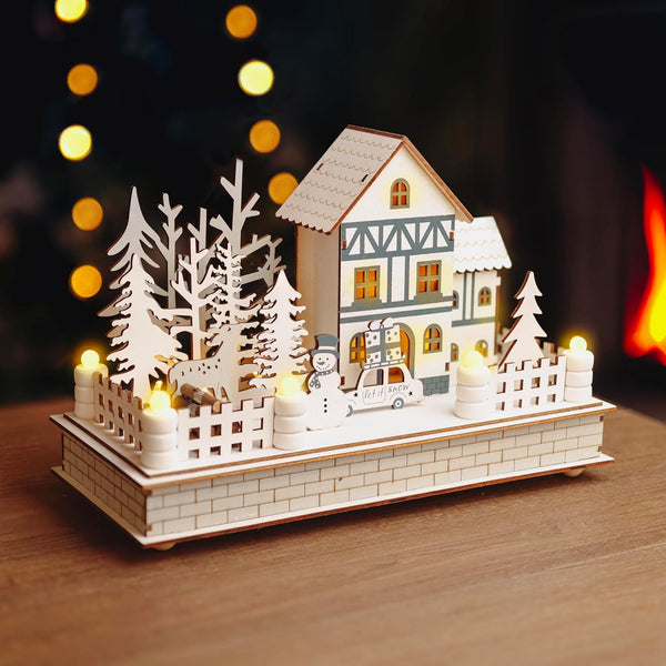 Decorative wooden house with trees and a snowman on a wooden surface with blurred lights in the background.