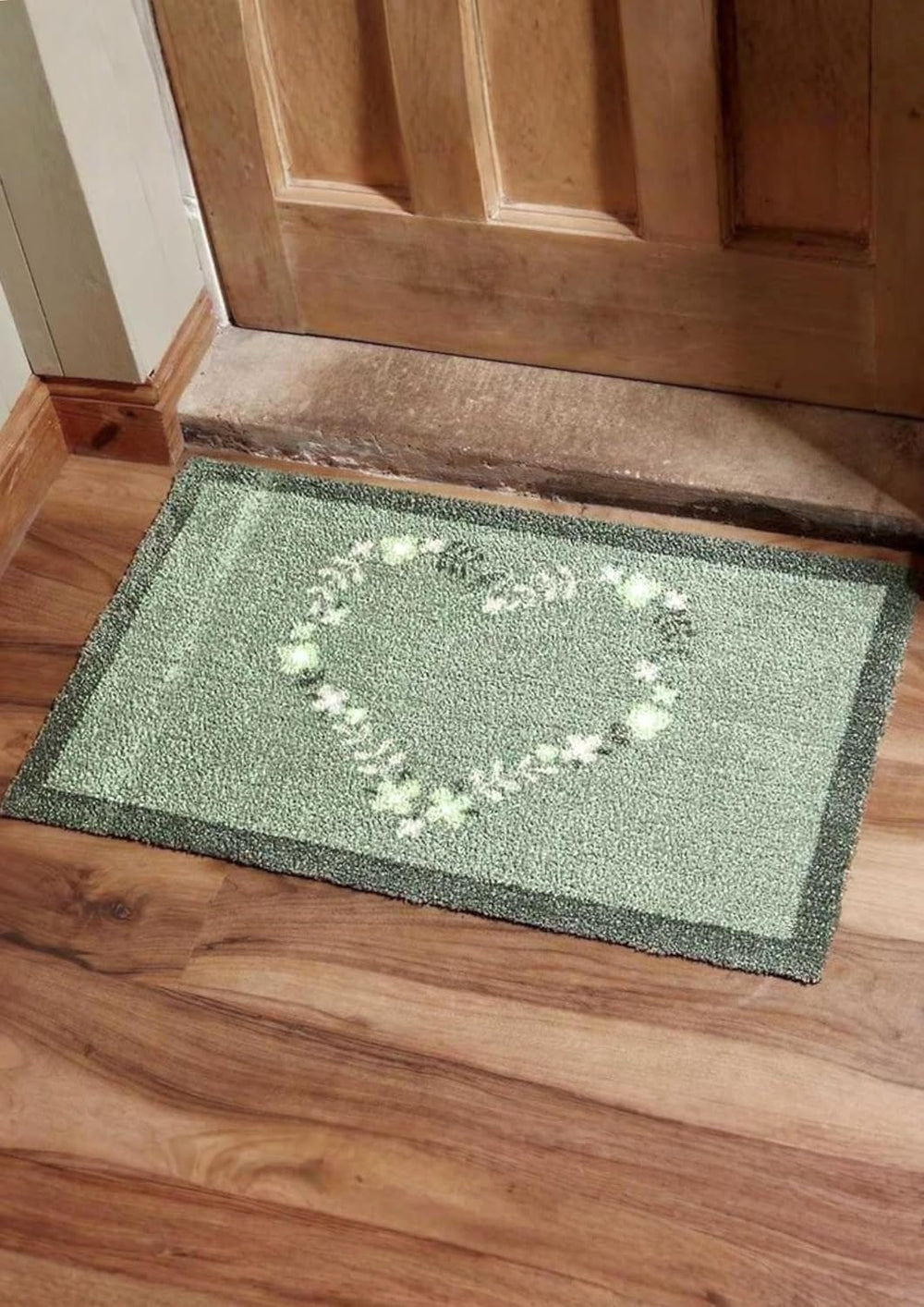Green doormat with a heart design on a wooden floor in front of a wooden door.