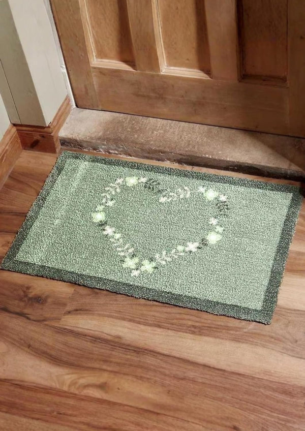 Green doormat with a heart design on a wooden floor in front of a wooden door.