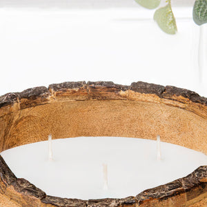 A close-up of a large wooden vessel candle with a natural tree-slice rim, filled with white wax and three visible wicks.