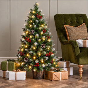 A decorated artificial Christmas tree stands in a living room setting next to an olive green armchair, with wrapped gift boxes placed at its base. The tree is green with white snow-dusted pinecones, white berries, and warm yellow fairy lights.