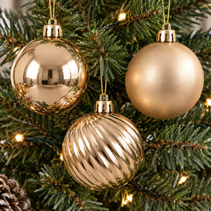 Three gold Christmas tree ornaments hanging on a green pine tree with warm white string lights. One ornament is highly reflective, one is matte, and one has a ribbed textured pattern.